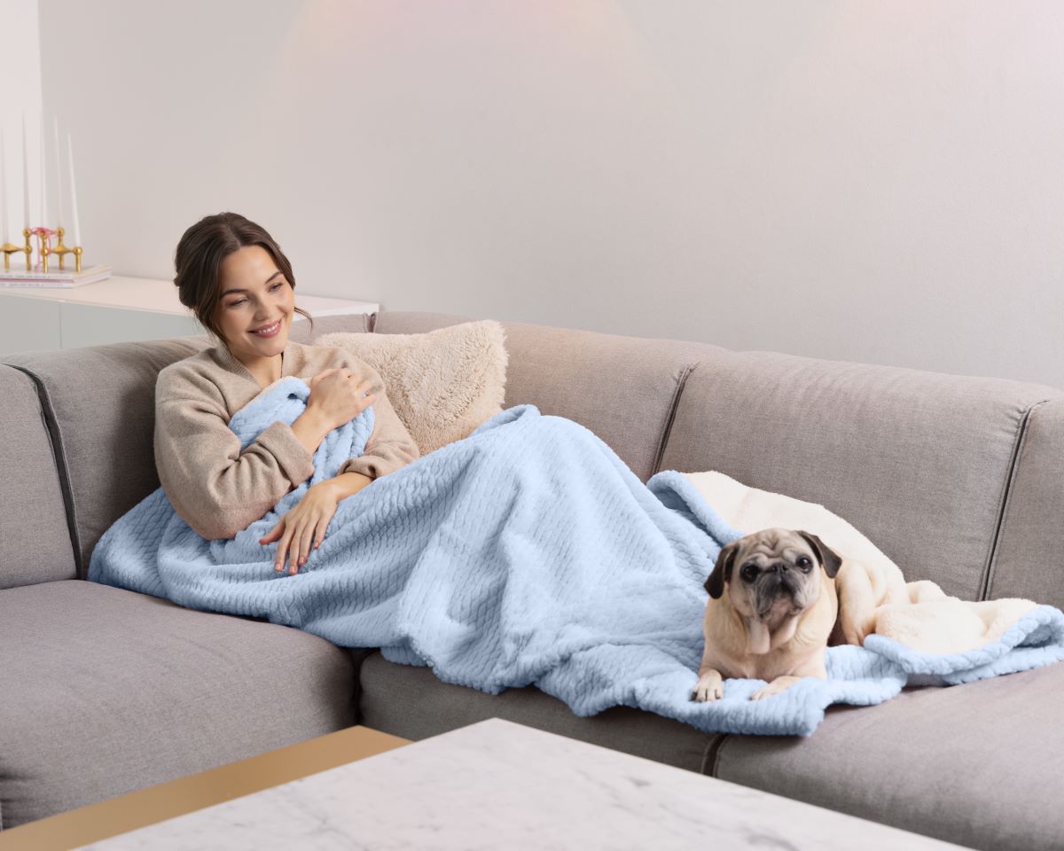 Frau entspannt auf dem Sofa mit einer hellblauen medisana Heizdecke, daneben liegt ein Hund.